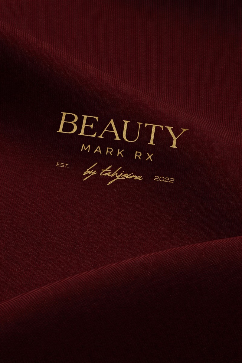 Beauty Mark RX logo in gold on a deep oxblood draped-fabric background — restrained and clinical-luxury.